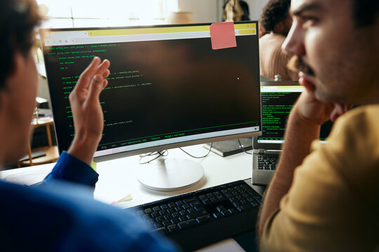 Male programmers discussing ideas with each other while coding on desktop PC in office