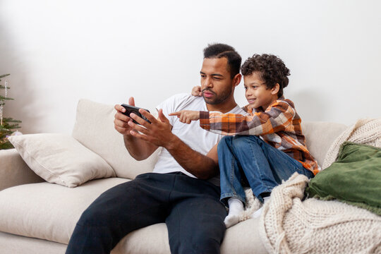 African American man and his son use smartphone at home on the couch and play video games online, teenager and his father look at the phone and are surprised