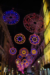 Colorful festive street christmas lights hanging above a city street at night, forming circular patterns and illuminating historic buildings, creating a vibrant holiday atmosphere with urban