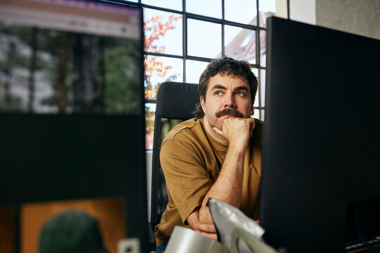 Contemplative male tech professional leaning on elbow while sitting at desk in startup office
