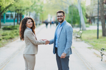 Obraz premium Professional man and woman in suits shaking hands in a public park, representing a business deal, academic partnership, or formal agreement outdoors.