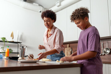 Fototapeta premium African-American woman and her son prepare chocolate butter and banana sandwiches in the kitchen, while teenager helps his mother prepare breakfast at home.