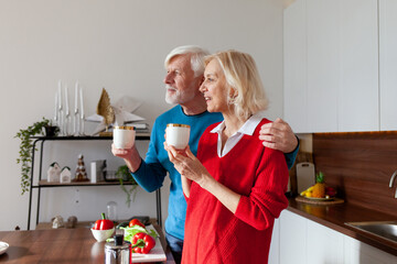 elderly couple of seniors drink coffee in the kitchen and smile, old woman and man hold cups of tea and hug at home