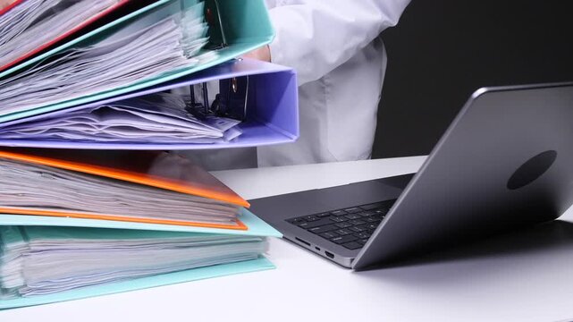 Medical professional in white lab coat balancing a towering, colorful stack of ring binders and paperwork beside an open laptop, illustrating administrative overload and paperwork burden