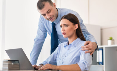 Fototapeta premium Sexual Harassment At Work. Businessman Sexually Harassing Employee Woman Touching Her Shoulder Working At Laptop Sitting In Office.