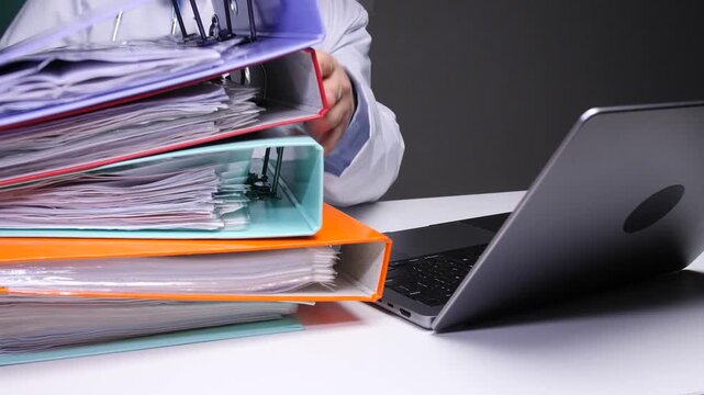 Doctor sitting at desk dealing with a large stack of patient files, binders, and medical documentation, representing the burden of healthcare bureaucracy