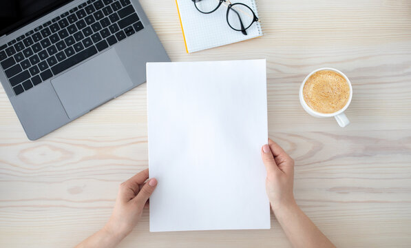 To do list or organizing plans. Woman holding white empty sheet of paper, sitting at desk with laptop computer and cup of coffee, top view, flat lay