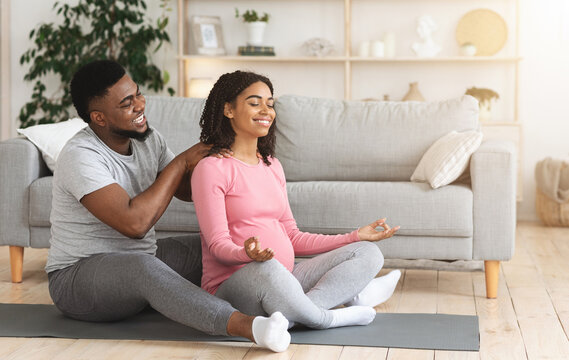 Loving African American husband massaging his meditating pregnant wife, sitting on floor at living room, exercising together, copy space