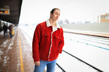 Obraz premium Young man in red jacket waiting on snowy train station platform during winter.