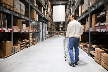 Rear view of person pushing shopping cart in large warehouse store aisle with tall shelves full of boxes. © Kyrylo