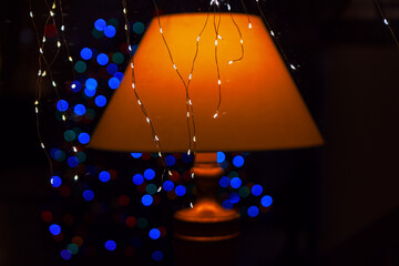 Cozy orange lampshade glows warmly beside delicate string lights, casting soft reflections. Behind it, blurred blue and red festive lights create a dreamy, atmospheric contrast