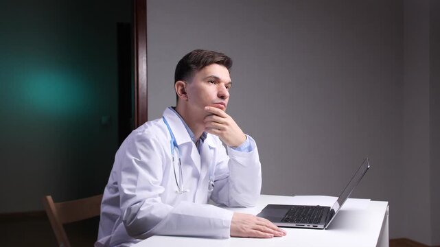 Male doctor in gown and stethoscope sitting at a desk with laptop, studying patient data with a thoughtful, concentrated expression as he analyzes diagnoses and treatment options