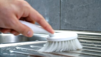 Hand holding a white plastic brush and scrubbing a metal kitchen sink with soap bubbles in a bright domestic setting