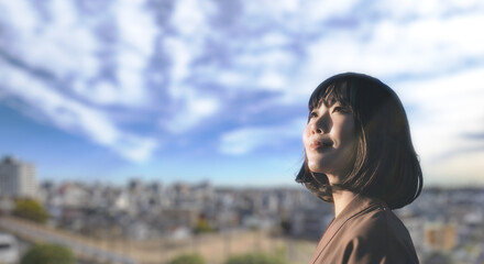 上を見上げ微笑む明るい表情の日本人女性の顔　前向きでポジティブ希望に満ちた表情　就職・新生活・旅行・リラックス・入学のイメージ　青空と街の背景