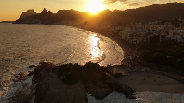 Drone descends while flying over Arpoador, capturing Ipanema Beach, ocean waves, crowded beach areas, and coastal buildings as the sun sets over Rio de Janeiro