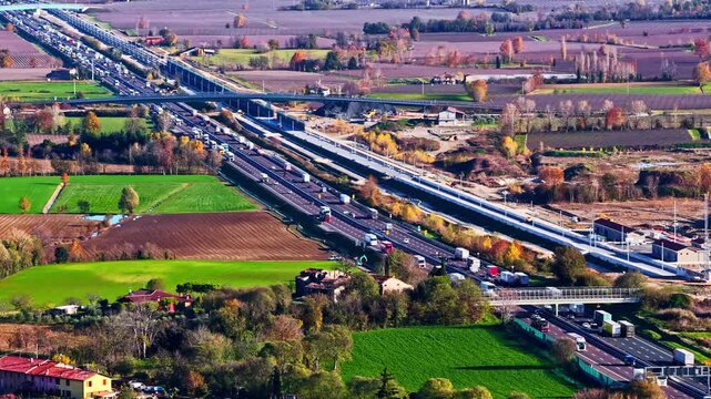 Aerial view of heavy traffic flowing on a multilane highway near Lago di Garda, surrounded by farmland, fields, and rural autumn landscapes on a clear bright day.