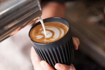 Barista pouring milk to create latte art in a black reusable coffee cup © n3moh3ro
