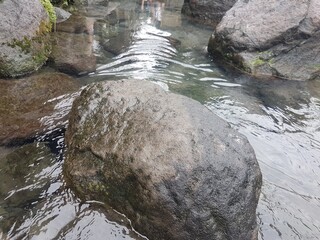 Large dark boulder partially submerged in flowing water. Wet, rough stone texture with water rippling around it. Natural river