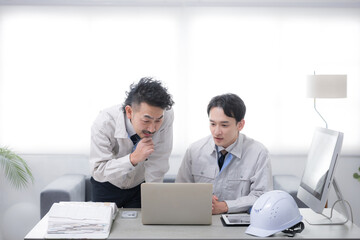 Obraz premium パソコンを見ながら話す作業着のビジネスマン2人 製造業などのイメージ 考える悩む顎に手を当てるポーズ