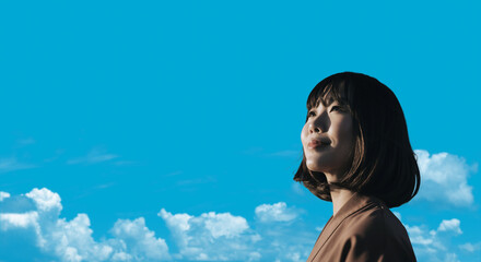 春・上を見上げ微笑む明るい表情の日本人女性の顔　前向きでポジティブ希望に満ちた表情　就職・新生活・旅行・リラックス・入学のイメージ　青空と雲の背景