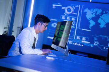 Young man coding in a modern office with blue lighting and data visualization on the wall