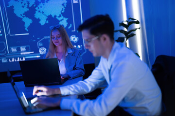 Two professionals collaborating on laptops in a modern workspace with a digital world map in the background at night
