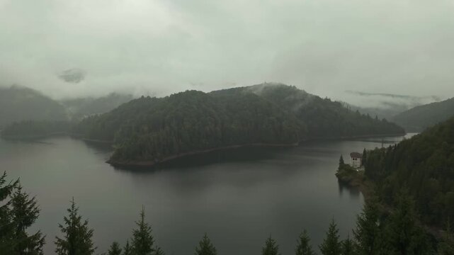 A mysterious view of Barajul Drăgan in Lunca Vişagului, Romania, featuring a serene reservoir surrounded by misty hills, lush greenery, and overcast skies on a rainy autumn day.