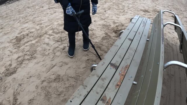 Person using a metal detector near a weathered beach bench, scanning the sand beside the seating area for lost items on a cold coastal day.