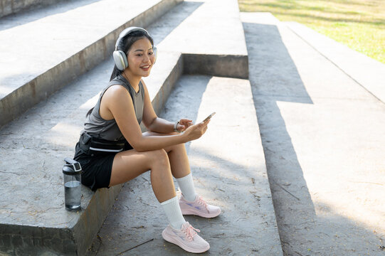 Smiling asian woman in sportswear listening to music in headphone sitting on concrete stairs in park