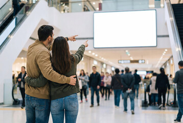 Couple embracing and pointing at a large blank billboard in a busy shopping mall
