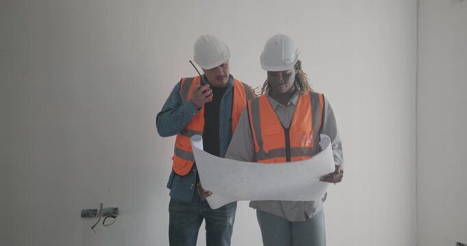 Engineer discuss blueprint at construction site, making decisions for architectural plan on upper floor. Both are dressed in safety gear. Collaborative approach to ensure project accuracy and progress