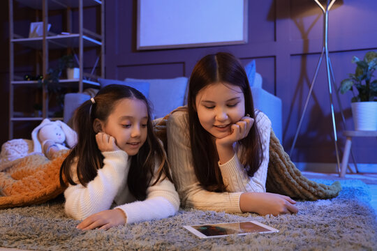 Cute Asian sisters with plaid watching cartoons on tablet computer at home in evening - Powered by Adobe