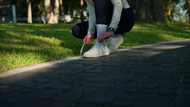 Close-up of a female runner stopping on a park path to tie her white sneakers. Active woman preparing for her morning workout routine
