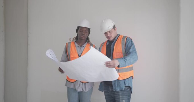 Engineer discuss blueprint at construction site, making decisions for architectural plan on upper floor. Both are dressed in safety gear. Collaborative approach to ensure project accuracy and progress