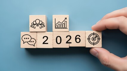 Hand arranging wooden blocks with icons representing business growth, teamwork, and targets, forming the year 2026 for future planning concepts.
