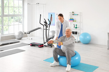 Fototapeta premium Female therapist and senior man with stretching band on fitball in rehabilitation center