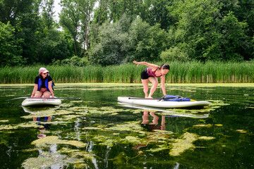 Two women have fun testing their balance on stand up paddle boards in an algae-filled pond on the Toronto Islands, with one playfully wobbling while her friend watches on a summer day.