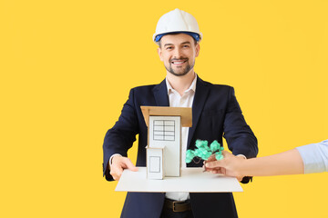 Male architect with 3D house model and female hand putting tree on yellow background