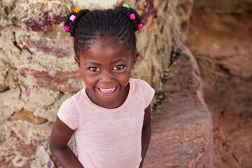 village happy african girl child profile portrait, cornrows braids dreadlocks hairstyle, big toothy smile, outdoors in the nature ,stone wall behind
