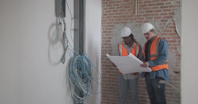 Engineer discuss blueprint at construction site, making decisions for architectural plan on upper floor. Both are dressed in safety gear. Collaborative approach to ensure project accuracy and progress