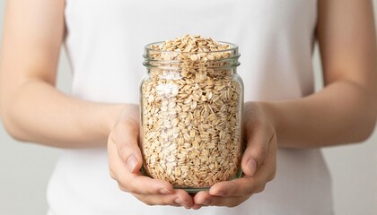 Hands holding a glass jar filled with rolled oats against a neutral background, suggesting healthy pantry storage and breakfast ingredients