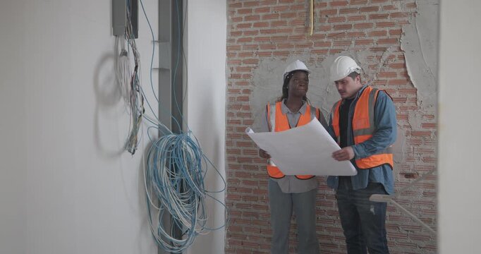 Engineer discuss blueprint at construction site, making decisions for architectural plan on upper floor. Both are dressed in safety gear. Collaborative approach to ensure project accuracy and progress