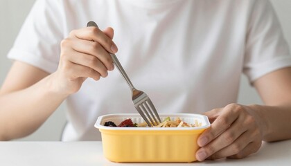 Person eating from a yellow plastic meal container with a fork at a table, close-up of hands and a casual packed lunch