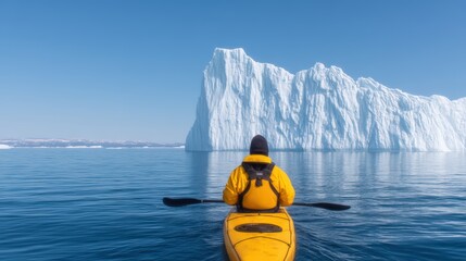 A kayaker in a yellow jacket paddles on calm waters, surrounded by towering icebergs under a clear blue sky.