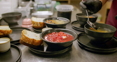 Catering cooking. Cafeteria lunch. Chef hand adding olives to hot steaming soup on kitchen table. Serving borsch in black bowl with bread.