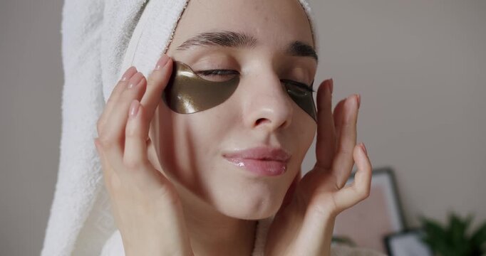 Beautiful woman in a bathrobe and a towel on head with healthy skin laughs joyfully applies brown patches to reduce eye bags. close up. Portrait Of Female Moisturizing Under Eye Skin Using Patches.