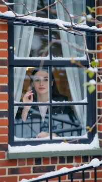 A young woman gazing thoughtfully through a snow-covered window, surrounded by the warmth of her cozy indoor setting