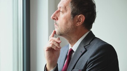 Mature businessman in suit standing by window with hand on chin, looking thoughtful and worried about work or decision. 4K.