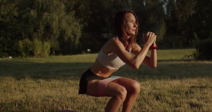 Athletic woman has workout and doing squat exercise with fitness resistance band outdoors. Young woman with slim body doing fitness exercises and squatting with resistance band outdoors.