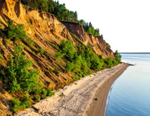 A coastal landscape features a high sand cliff, a sandy beach, and calm water reflecting soft sunlight. Trees dot the cliff
